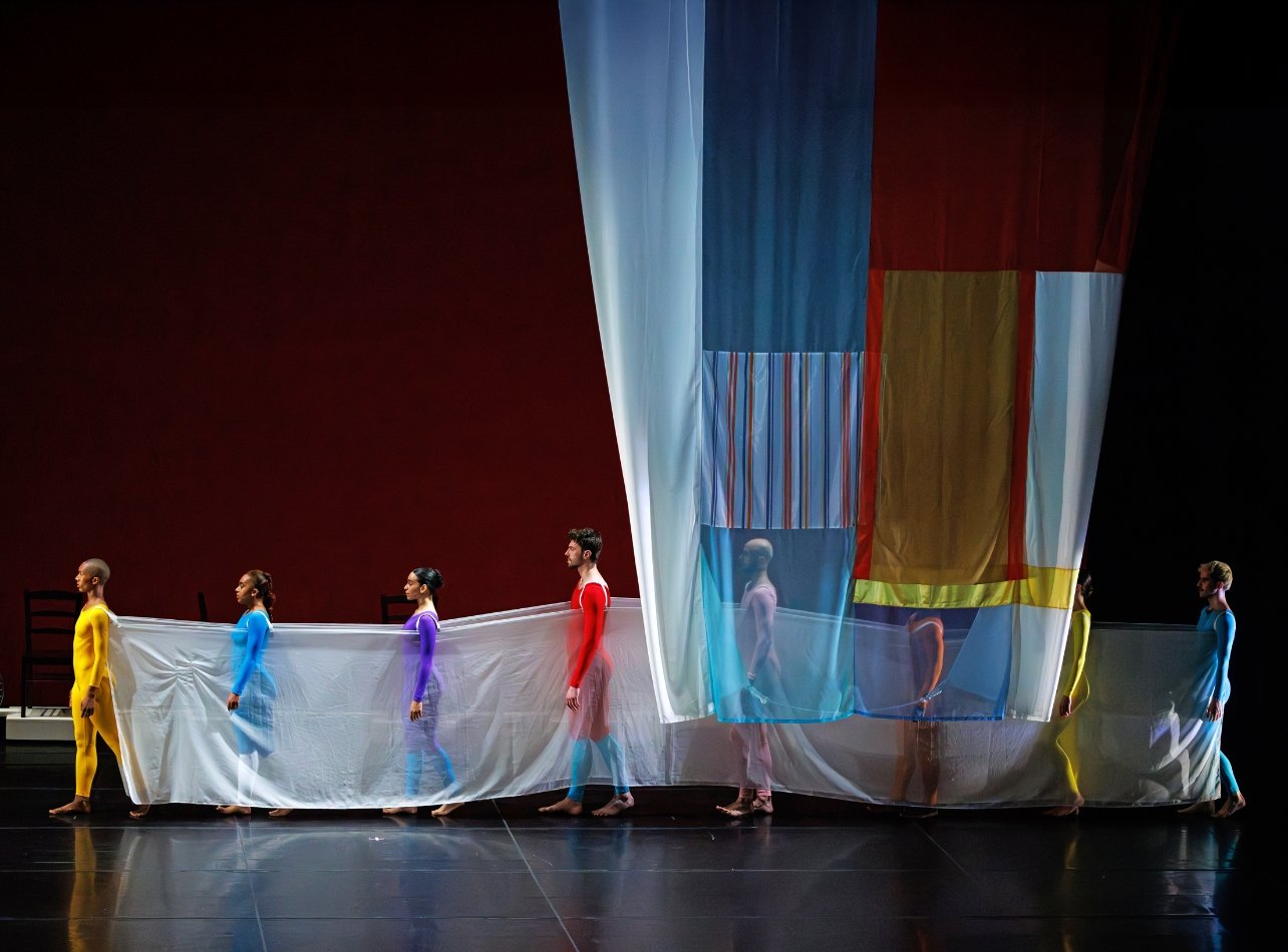 A choreographed dance festival with artists on stage dressed in colorful bodysuits carrying a cloth