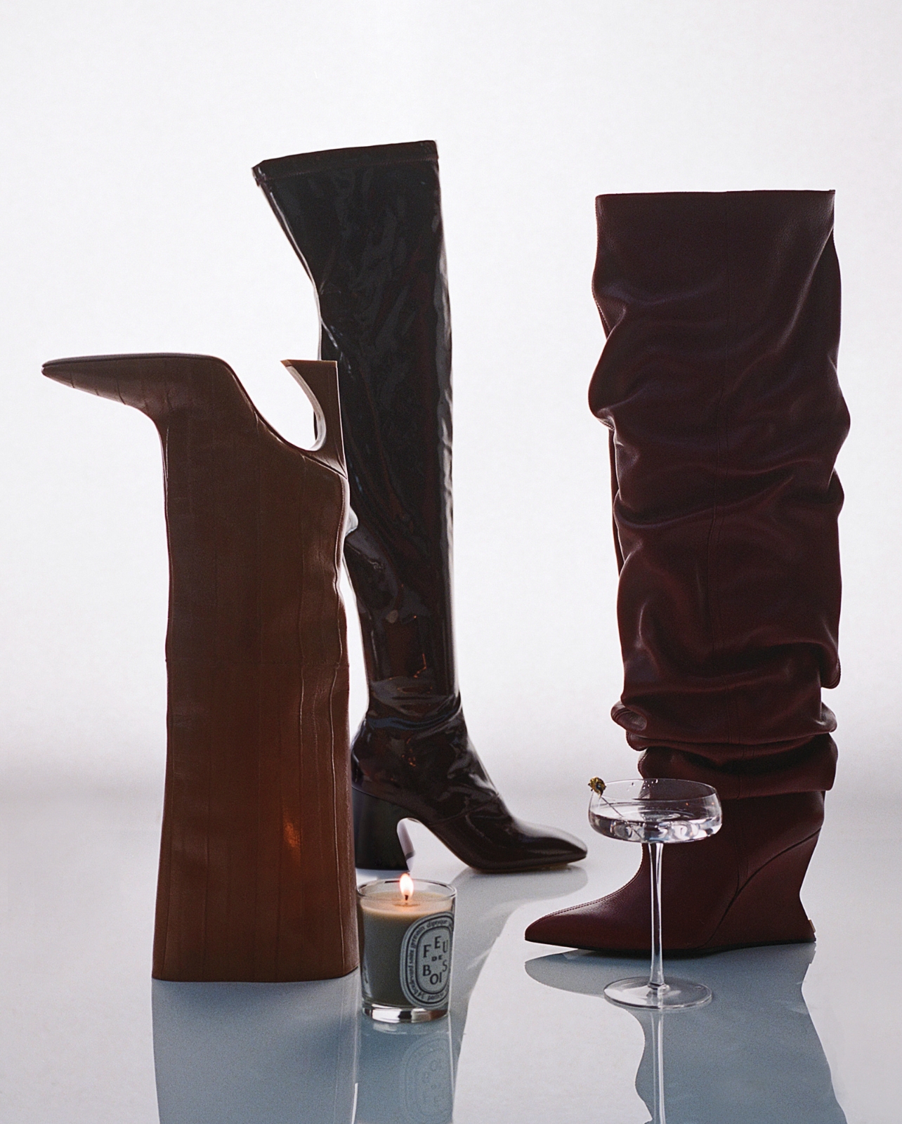 A still life photo of tall designer boots, a candle, and a cocktail glass