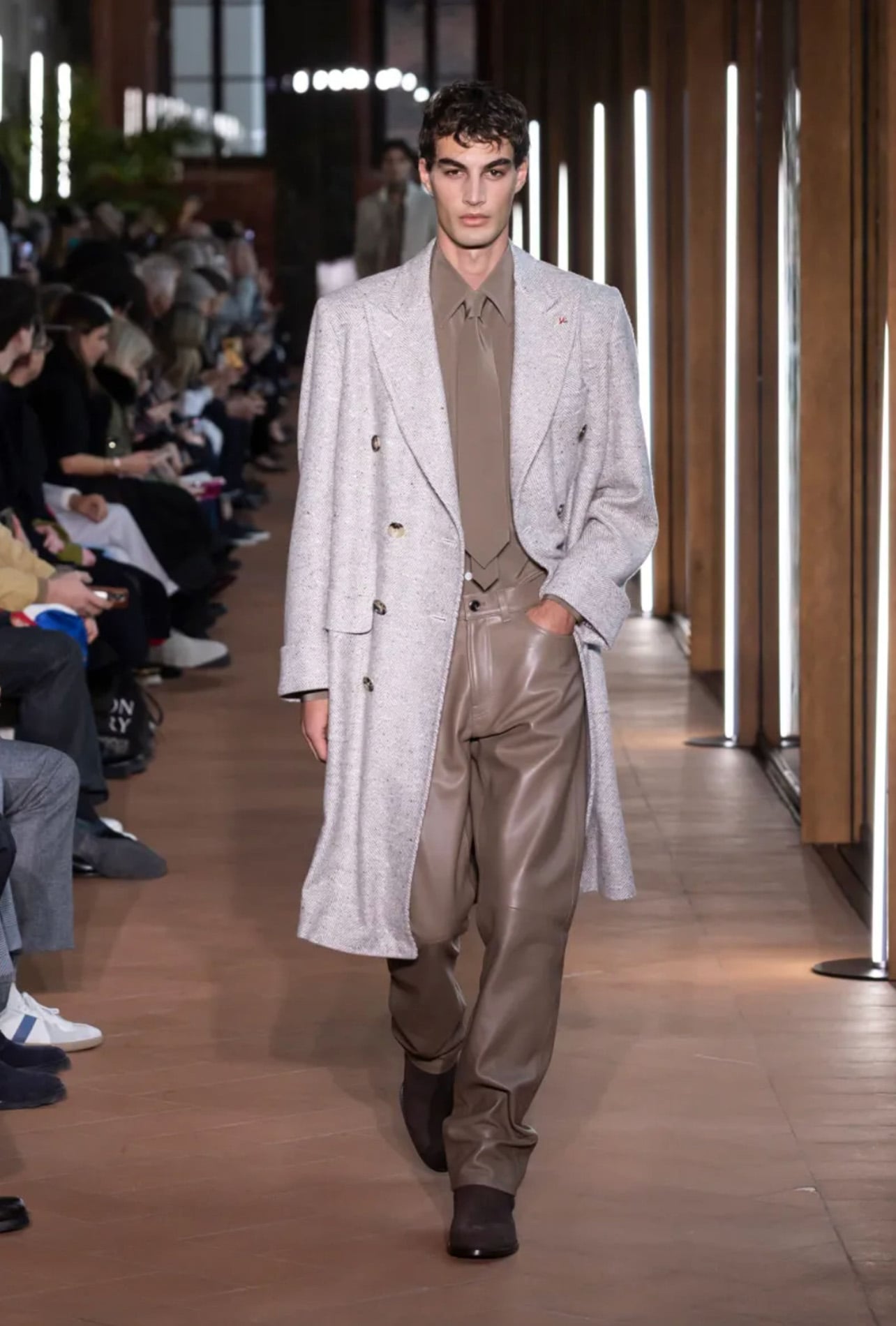 Isaia male model on runway wearing beige leather pants, beige button down and tie with long grey coat