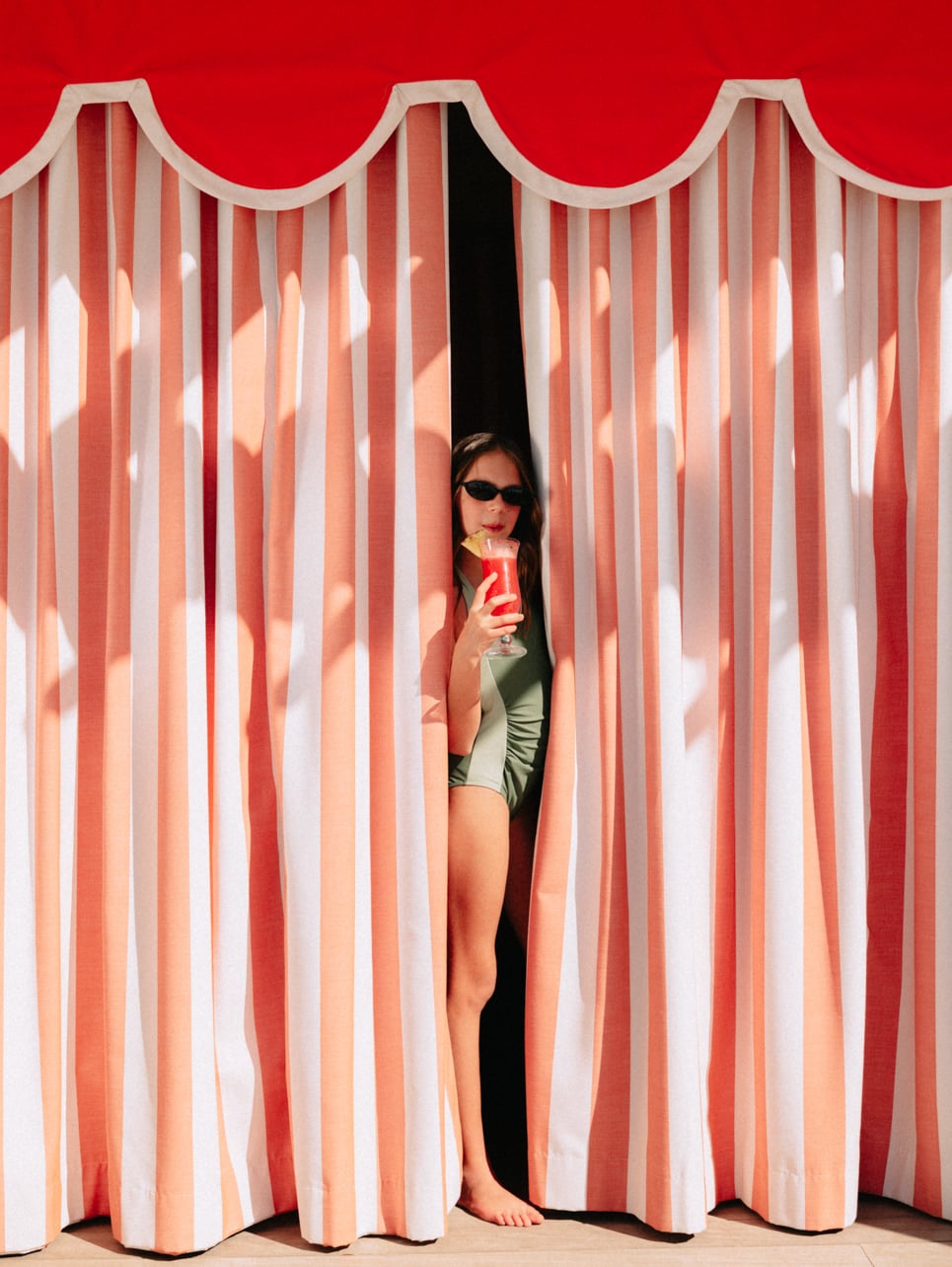A girl peeking through a curtain wearing a bathing suit and sunglasses