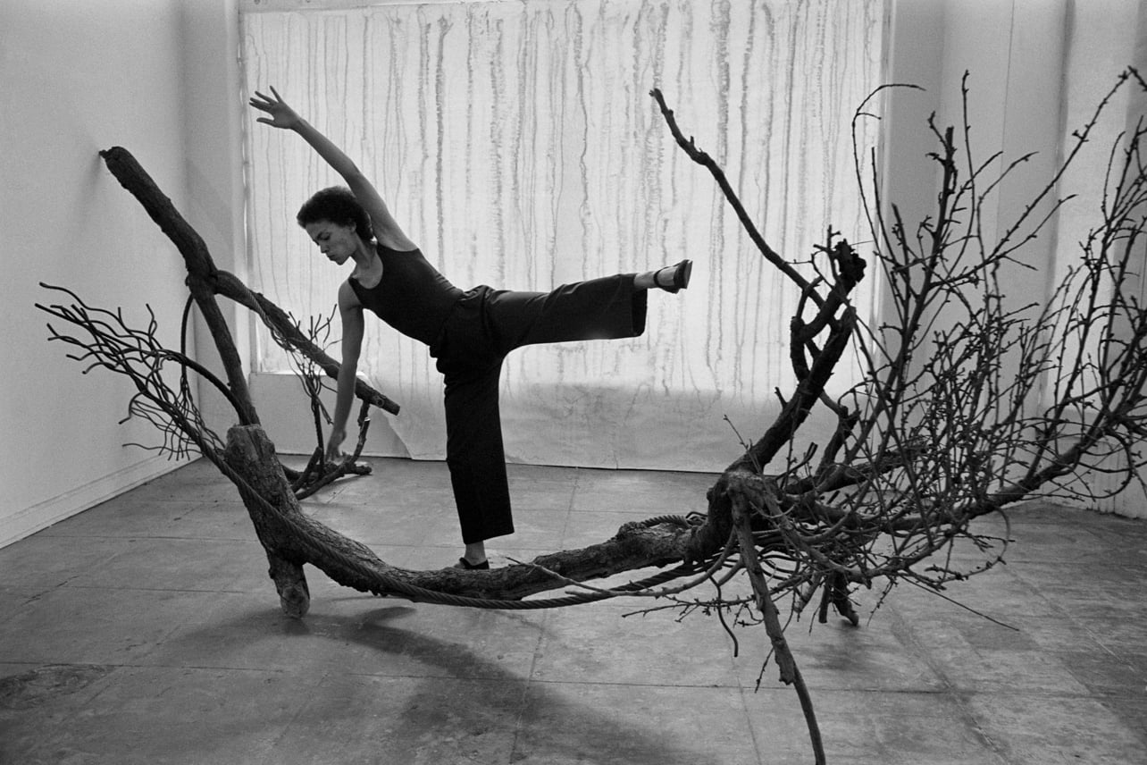 A black and white photograph featuring a woman interacting with tree branches by Maren Hassinger