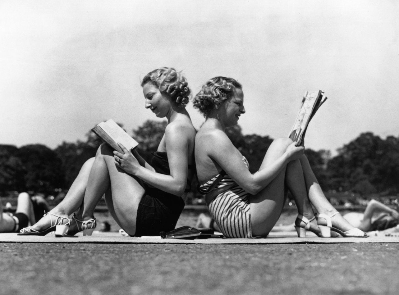 A black-and-white archival photograph of two women sitting back-to-back while reading outdoors