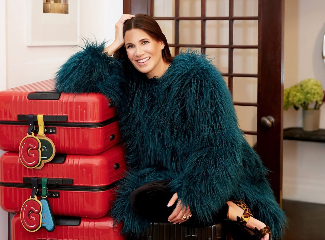 A portrait of Elizabeth Saltzman in a feathered jacket leaning on a stack of red suitcases