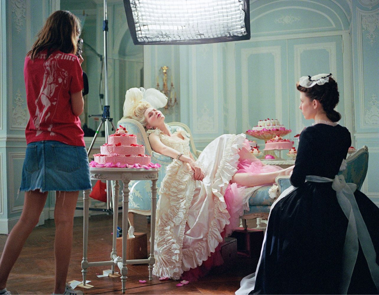 A photograph of Kirsten Dunst wearing a long dress surrounded by pink cakes