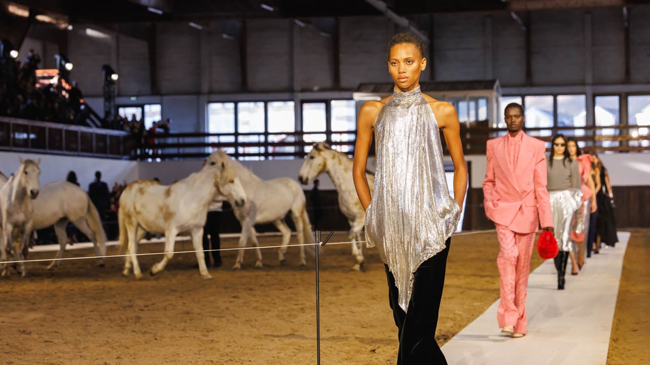 A close-up photo of runway models at the Stella McCartney show wearing silver, black, and pink pieces at a riding hall with horses